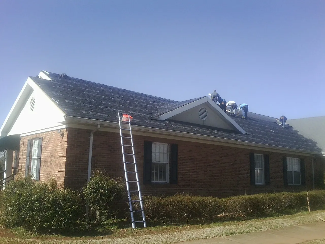 Crew tearing shingles off a brick ranch roof with a ladder set — Emergency Roof Repair work by our crew in Belmond