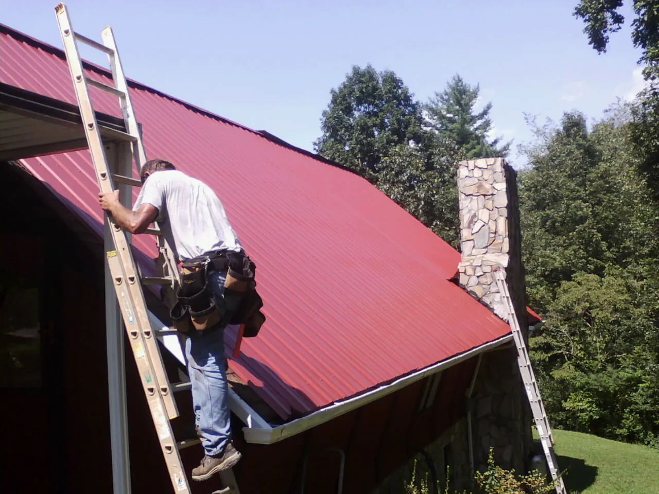 Roofer on a ladder beside a red metal roof and stone chimney — Soffit & Fascia Repair by Roofing Brothers in Belmond